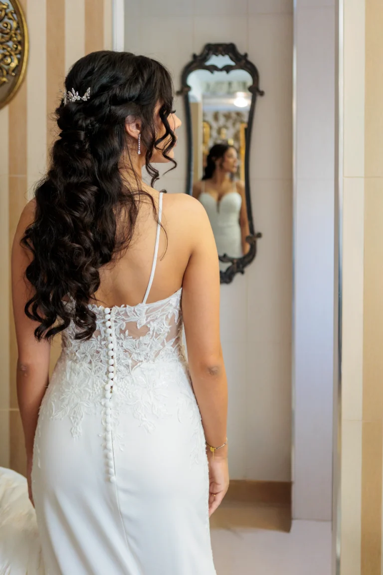 Espalda de novia con vestido blanco de encaje y botones forrados, mirando su reflejo en un espejo antiguo durante los preparativos de boda.
