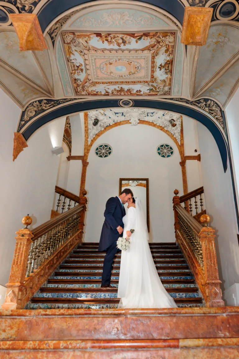 Novios abrazados en las escaleras de una iglesia decorada en Jaén con techo artístico y arquitectura elegante.