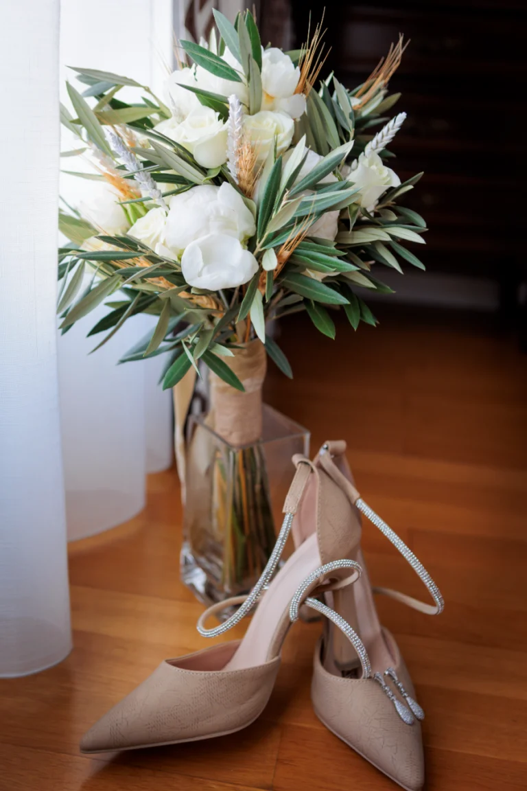 Ramo de novia con rosas blancas y hojas de olivo junto a zapatos nude de tacón sobre suelo de madera.