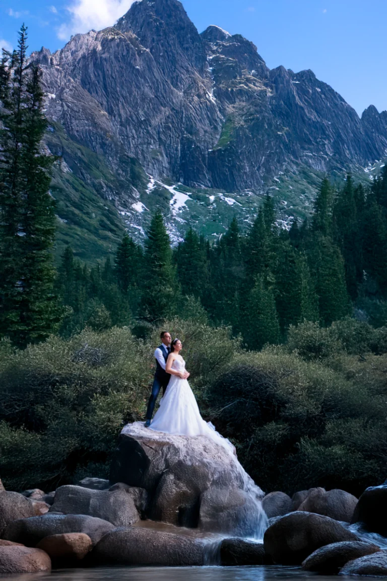 Novios sobre una roca junto a un río con montañas al fondo durante su reportaje de boda en exterior