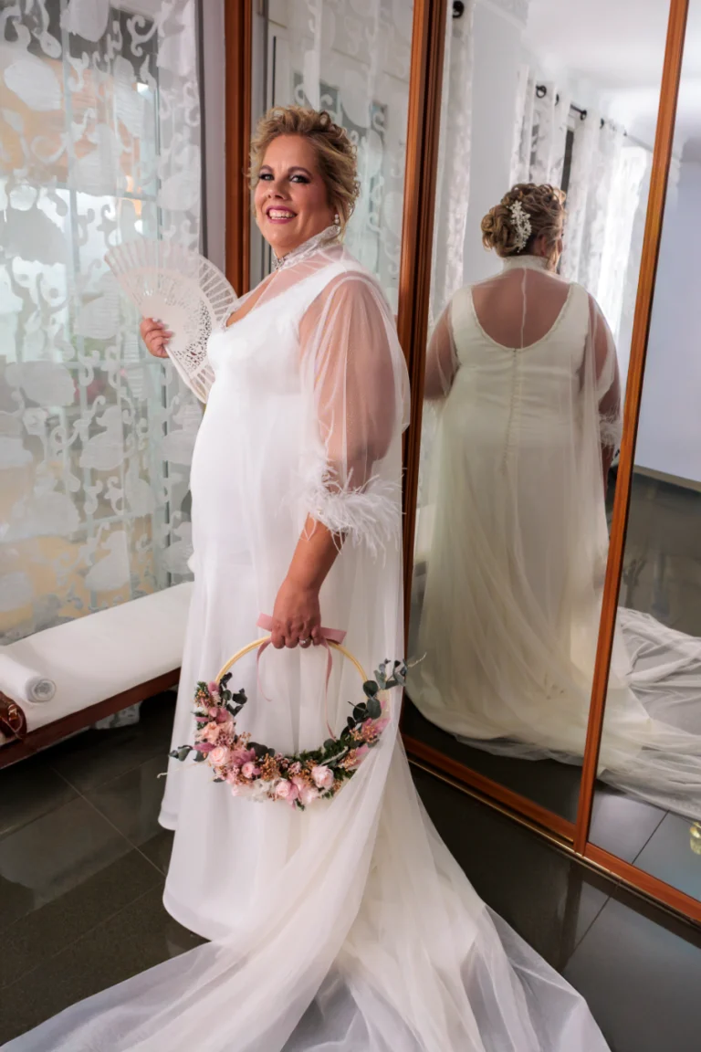 Novia de espaldas junto al espejo sosteniendo un tocado floral durante los preparativos de boda en casa.